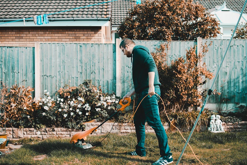 A man with short hair, wearing glasses, a dark hooded sweatshirt, and jeans, is performing lawn maintenance in a garden. He is using an electric lawn trimmer to trim the grass along the edge of a lush
