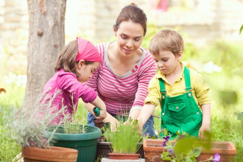 A woman with dark brown hair, wearing a pink and white striped long-sleeve shirt, is engaged in gardening alongside two young children in a lush outdoor garden. One child, a girl in a bright pink hat and matching jacket, is reaching into a gardening pot, while the other, a boy dressed in a yellow shirt and green overalls, is inspecting the soil or plants in a rectangular terracotta planter. The garden features a variety of potted plants on a wooden table, with green foliage and blooming flowers visible in the background. A large tree trunk is on the left, providing partial shade, and the surrounding environment is vibrant with natural light, suggesting a clear, sunny day. The scene highlights family participation in outdoor gardening activities, with the garden area comprising grassy patches, flower beds, and well-maintained planting containers, aligning with local gardening services like those offered by Gardener Brompton in the Brompton area of London, UK.